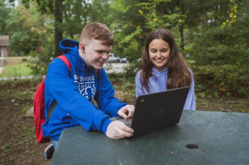 Exeter-West Greenwich Buddy Pair Young man in blue hoodie and young woman in purple hoodie smile at a computer while sitting at a table outside